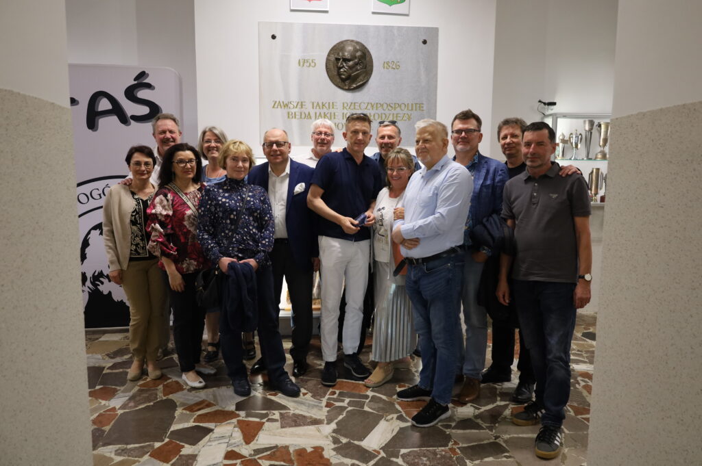 Zdjęcie przedstawia grupę 17 dorosłych osób (9 mężczyzn i 8 kobiet), pozujących razem do pamiątkowego zdjęcia w jasnym, przestronnym wnętrzu. Wszyscy stoją blisko siebie, zwróceni przodem do obiektywu aparatu. Panuje atmosfera koleżeńska i serdeczna, wiele osób się uśmiecha. Większość postaci ma luźno opuszczone ręce, niektóre obejmują się wzajemnie lub trzymają się lekko za ramiona. Lokalizacja i tło: Zdjęcie wykonane jest w holu szkolnym lub korytarzu z jasnym oświetleniem sztucznym (brak okien w kadrze). Na ścianie za grupą znajduje się duża, pionowa tablica pamiątkowa z białego marmuru z wizerunkiem twarzy w formie wypukłego medalu (profil starszego mężczyzny z poważnym wyrazem twarzy). Nad medalionem wypisane są daty 1755–1826, a poniżej widnieje złoty napis z cytatem: „Zawsze takie Rzeczypospolite będą, jakie ich młodzieży chowanie.” Cytat przypisywany Stanisławowi Staszicowi, odnoszący się do roli wychowania młodzieży w kształtowaniu państwa. Powyżej tablicy widać dwa herby, jeden czerwony, drugi zielony (częściowo ucięte w kadrze). Po prawej stronie znajduje się oszklona gablota z wieloma pucharami i trofeami sportowymi, rozświetlona od wewnątrz. Po lewej stronie częściowo widoczna grafika ścienna z dużym, czarnym napisem: „TAŚ” oraz fragmentem logo szkolnego z konturem profilu mężczyzny. Podłoga i otoczenie: Podłoga składa się z wielokolorowych, nieregularnych płytek z wzorem mozaiki (w odcieniach brązu, bieli, beżu i czerni). Po bokach zdjęcia znajdują się dwie kolumny lub filary, wyłożone tynkiem w odcieniu jasnoszarym z fakturą ziarnistą. Wygląd uczestników: Ubrania są codzienne lub półformalne: spodnie, jeansy, swetry, koszule, marynarki, bluzki w kwiaty, tuniki. Wśród kobiet widoczne są torebki na ramieniu i okulary, zarówno korekcyjne jak i przeciwsłoneczne (na głowie). W centrum zdjęcia mężczyzna w ciemnogranatowej koszulce polo i białych spodniach trzyma niewielki przedmiot (prawdopodobnie okulary lub etui) i otoczony jest innymi osobami, co może sugerować, że jest ważną postacią dla tej grupy. Wszyscy są w wieku około 50–60 lat, co sugeruje, że może to być spotkanie absolwentów szkoły po latach. Kontekst: Zdjęcie ma charakter wspomnieniowy, nieformalny i serdeczny. Sugeruje ponowne spotkanie znajomych ze szkoły, np. z okazji rocznicy ukończenia liceum. Lokalizacja, cytat i elementy szkolne w tle jednoznacznie wskazują na miejsce z historią edukacyjną i wychowawczą.