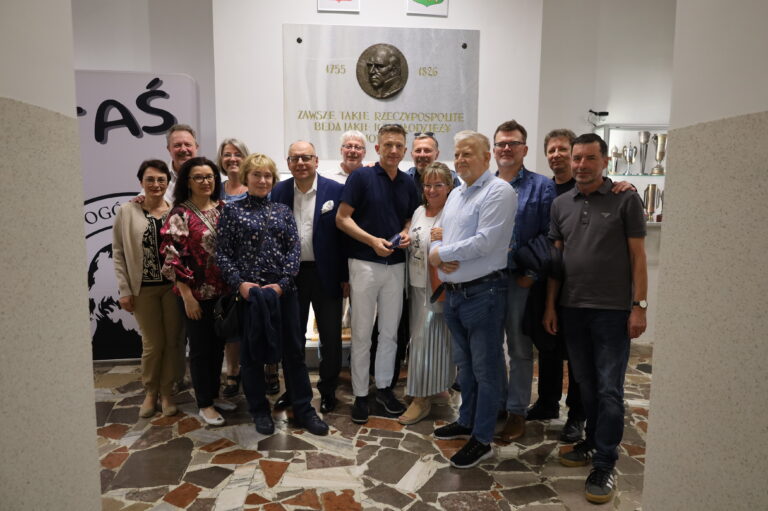 Zdjęcie przedstawia grupę 17 dorosłych osób (9 mężczyzn i 8 kobiet), pozujących razem do pamiątkowego zdjęcia w jasnym, przestronnym wnętrzu. Wszyscy stoją blisko siebie, zwróceni przodem do obiektywu aparatu. Panuje atmosfera koleżeńska i serdeczna, wiele osób się uśmiecha. Większość postaci ma luźno opuszczone ręce, niektóre obejmują się wzajemnie lub trzymają się lekko za ramiona. Lokalizacja i tło: Zdjęcie wykonane jest w holu szkolnym lub korytarzu z jasnym oświetleniem sztucznym (brak okien w kadrze). Na ścianie za grupą znajduje się duża, pionowa tablica pamiątkowa z białego marmuru z wizerunkiem twarzy w formie wypukłego medalu (profil starszego mężczyzny z poważnym wyrazem twarzy). Nad medalionem wypisane są daty 1755–1826, a poniżej widnieje złoty napis z cytatem: „Zawsze takie Rzeczypospolite będą, jakie ich młodzieży chowanie.” Cytat przypisywany Stanisławowi Staszicowi, odnoszący się do roli wychowania młodzieży w kształtowaniu państwa. Powyżej tablicy widać dwa herby, jeden czerwony, drugi zielony (częściowo ucięte w kadrze). Po prawej stronie znajduje się oszklona gablota z wieloma pucharami i trofeami sportowymi, rozświetlona od wewnątrz. Po lewej stronie częściowo widoczna grafika ścienna z dużym, czarnym napisem: „TAŚ” oraz fragmentem logo szkolnego z konturem profilu mężczyzny. Podłoga i otoczenie: Podłoga składa się z wielokolorowych, nieregularnych płytek z wzorem mozaiki (w odcieniach brązu, bieli, beżu i czerni). Po bokach zdjęcia znajdują się dwie kolumny lub filary, wyłożone tynkiem w odcieniu jasnoszarym z fakturą ziarnistą. Wygląd uczestników: Ubrania są codzienne lub półformalne: spodnie, jeansy, swetry, koszule, marynarki, bluzki w kwiaty, tuniki. Wśród kobiet widoczne są torebki na ramieniu i okulary, zarówno korekcyjne jak i przeciwsłoneczne (na głowie). W centrum zdjęcia mężczyzna w ciemnogranatowej koszulce polo i białych spodniach trzyma niewielki przedmiot (prawdopodobnie okulary lub etui) i otoczony jest innymi osobami, co może sugerować, że jest ważną postacią dla tej grupy. Wszyscy są w wieku około 50–60 lat, co sugeruje, że może to być spotkanie absolwentów szkoły po latach. Kontekst: Zdjęcie ma charakter wspomnieniowy, nieformalny i serdeczny. Sugeruje ponowne spotkanie znajomych ze szkoły, np. z okazji rocznicy ukończenia liceum. Lokalizacja, cytat i elementy szkolne w tle jednoznacznie wskazują na miejsce z historią edukacyjną i wychowawczą.