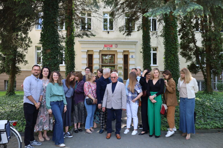 Na zdjęciu przed wejściem do budynku szkoły średniej (styl klasycystyczny, elewacja jasnożółta z białymi akcentami wokół okien i drzwi) stoi grupa 19 osób – mężczyzn i kobiet, w różnym wieku, głównie dorosłych, ustawionych nieregularnie. W centrum stoi starszy mężczyzna w jasnej marynarce, granatowej koszuli i okularach przeciwsłonecznych. Pozostałe osoby są w bardziej swobodnych pozach – niektóre patrzą w obiektyw, inne rozmawiają ze sobą, śmieją się, odwracają wzrok, poprawiają włosy – zdjęcie ma charakter nieformalny, jakby wykonane spontanicznie, w przerwie spotkania. W tle widać wejście główne do szkoły z dwuskrzydłowymi drewnianymi drzwiami, nad którymi znajdują się: Czerwona tablica z napisem informującym o placówce edukacyjnej, Godło Polski – biały orzeł na czerwonym tle. Otoczenie to zieleń – po bokach drzewa z gęsto porośniętymi pniami bluszczem oraz nisko przycięte krzewy. Grupa stoi na chodniku z szarej kostki brukowej.