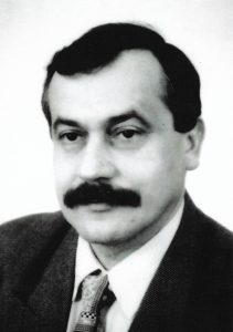 Czarno-białe zdjęcie portretowe przedstawia mężczyznę w średnim wieku, ujętego od klatki piersiowej wzwyż, w pozycji frontalnej. Mężczyzna patrzy prosto w obiektyw z poważnym, spokojnym wyrazem twarzy. Ujęcie jest wyraźne i ostre, co pozwala na łatwe rozpoznanie szczegółów. Twarz mężczyzny ma owalny kształt z gładkim czołem i wyraźnie zarysowanymi brwiami. Jego spojrzenie jest skupione, oczy średniej wielkości, osadzone symetrycznie. Ma charakterystyczny, gęsty i ciemny wąs, który zajmuje całą szerokość nad górną wargą. Jego nos jest prosty, średniej długości, a usta zamknięte, neutralnie ułożone. Policzki są pełne, podbródek łagodnie zaokrąglony. Włosy mężczyzny są ciemne, gęste, zaczesane na bok. Linia włosów jest łagodna, z lekkim cofnięciem w okolicach skroni, ale bez oznak łysienia. Ma krótkie bokobrody, które płynnie łączą się z włosami. Mężczyzna ubrany jest elegancko: ma na sobie jasną koszulę z kołnierzykiem, krawat o geometrycznym wzorze (prawdopodobnie kratka lub delikatne paski) oraz ciemną marynarkę o fakturze sugerującej grubszą tkaninę – może to być wełna lub tweed. Tło zdjęcia jest jednolite i jasne, prawdopodobnie białe lub bardzo jasnoszare, co nadaje zdjęciu czysty, formalny charakter. Styl fotografii oraz ubiór sugerują, że zdjęcie pochodzi z końca XX wieku, być może z lat 80. lub 90. Zdjęcie ma typowy charakter fotografii urzędowej lub akademickiej – mogło być wykonane na potrzeby dokumentów, publikacji naukowej lub materiałów promocyjnych. Całość kompozycji sprawia wrażenie profesjonalizmu i powagi.