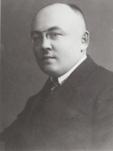 Czarno-białe zdjęcie portretowe przedstawia mężczyznę w średnim wieku, ujętego w pozycji siedzącej, zwróconego delikatnie w lewo, ale patrzącego bezpośrednio w stronę obiektywu. Ujęcie obejmuje górną część jego sylwetki – od klatki piersiowej wzwyż. Mężczyzna ma owalną, pełną twarz z łagodnym, spokojnym wyrazem. Jego oczy są półprzymknięte, spojrzenie neutralne, usta zamknięte, bez wyraźnego uśmiechu. Głowa jest niemal całkowicie łysa na górze – widoczna jest gładka, wypolerowana powierzchnia czaszki. Włosy pozostały jedynie po bokach i z tyłu głowy – są krótkie i ciemne, starannie przystrzyżone. Mężczyzna nosi cienkie okulary z metalowymi oprawkami, które podkreślają jego intelektualny wizerunek. Nos jest prosty i dość szeroki, policzki pełne, a podbródek mocno zarysowany. Ubrany jest bardzo elegancko: ma na sobie ciemny garnitur, białą koszulę z klasycznym kołnierzykiem oraz krawat – wzorzysty, prawdopodobnie w ciemnych odcieniach, choć szczegóły wzoru nie są wyraźne ze względu na kontrast zdjęcia. Tło jest jednolite, ciemnoszare i całkowicie neutralne – pozbawione jakichkolwiek szczegółów czy elementów. Zdjęcie ma charakter formalny, najprawdopodobniej wykonane zostało w studio fotograficznym. Styl ubioru oraz ogólna estetyka zdjęcia sugerują, że fotografia pochodzi z pierwszej połowy XX wieku, być może z lat 20. lub 30. Postać na zdjęciu prezentuje się jako osoba poważna, zaufana, być może nauczyciel, naukowiec, urzędnik lub przedstawiciel zawodu wymagającego wysokiej kultury osobistej.