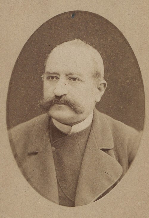 Fotografia przedstawia portret mężczyzny w średnim wieku lub starszego, wykonany w technice sepii (brązowo-szarej tonacji), prawdopodobnie z końca XIX lub początku XX wieku. Obraz ma kształt pionowego owalu, otoczonego beżowym tłem w kształcie prostokąta. Wygląd mężczyzny: Mężczyzna ma jasną, nieco przerzedzoną fryzurę – włosy są krótkie i zaczesane do tyłu. Skóra jasna, twarz okrągła, z wyraźnymi rysami starzenia, takimi jak delikatne zmarszczki i opadnięte powieki. Dominującym elementem twarzy są bardzo bujne, gęste wąsy typu „kierownica” – szerokie i zakręcone ku bokom, gęste, ciemniejsze od włosów. Brwi są również wyraźne, ale nie tak gęste jak wąsy. Ubiór: Mężczyzna ubrany jest formalnie, co wskazuje na status społeczny lub profesjonalny charakter zdjęcia. Ma na sobie ciemny surdut (płaszcz lub marynarka) z szerokimi klapami i widocznymi guzikami. Pod spodem nosi kamizelkę i biały kołnierzyk koszuli, zapięty wysoko pod szyją, co było typowe dla mody męskiej przełomu XIX i XX wieku. Tło i styl fotografii: Tło jest jednolite i rozmyte, co skupia uwagę wyłącznie na twarzy i sylwetce mężczyzny. Fotografia wykonana w stylu atelierowym – pozowane zdjęcie portretowe, bez uśmiechu, z lekkim spojrzeniem w lewą stronę (czyli w prawo z perspektywy widza). Całość wpisana jest w owalny kadr, typowy dla dawnych fotografii portretowych (tzw. „wizytówka” – carte de visite). Charakterystyka techniczna: Zdjęcie wygląda na odbitkę na kartonie fotograficznym, z lekkimi oznakami starzenia – widoczne są plamki, zacieki i drobne uszkodzenia papieru. Podsumowanie wizualne: To formalny portret starszego mężczyzny z charakterystycznymi, dużymi wąsami, ubrany w elegancki, klasyczny strój z przełomu wieków. Styl zdjęcia i ubiór wskazują, że był to człowiek prawdopodobnie należący do wyższych sfer społecznych lub pełniący funkcję publiczną, np. urzędnika, lekarza czy naukowca.