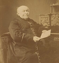 Portret studyjny starszego, łysiejącego mężczyzny w sutannie (duchownym stroju) siedzącego na fotelu. Zdjęcie jest sepia-tonowane (brązowawy odcień), wykonane w technice fotograficznej typowej dla końca XIX wieku. Opis szczegółowy: Mężczyzna siedzi na fotelu z dekoracyjnymi frędzlami po bokach, lekko bokiem do obiektywu. Jego twarz jest pełna, z łagodnym wyrazem – patrzy lekko na bok, jakby zamyślony. Ubrany jest w sutannę zapinaną na rząd guzików z przodu, z ozdobnym wykończeniem przy rękawach. W dłoniach trzyma złożony arkusz papieru lub list. W tle znajduje się zdobiony mebel – prawdopodobnie biurko lub sekretarzyk – z ozdobną kratką i tkaniną. Scena sfotografowana w atelier, z jednolitym tłem i miękkim światłem podkreślającym rysy twarzy.