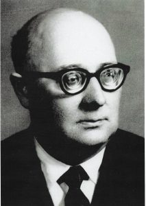 Czarno-białe zdjęcie portretowe przedstawia mężczyznę w średnim wieku lub starszego, ujętego od klatki piersiowej w górę. Mężczyzna patrzy lekko w prawo z powagą i skupieniem. Ma okrągłą twarz i wysokie czoło. Górna część jego głowy, zwłaszcza po bokach i z tyłu, jest wyraźnie łysa, z niewielką ilością włosów pozostających tylko na dolnej części czaszki, z tyłu głowy i nieco nad uszami. Skóra głowy jest gładka i lśniąca. Mężczyzna nosi grube, ciemne okulary z zaokrąglonymi, dość dużymi oprawkami. Szkła są grube, co powoduje lekkie zniekształcenie widocznych za nimi oczu. Oczy są szeroko otwarte, a spojrzenie wydaje się intensywne i skupione, skierowane nieco w bok, poza kadr. Nos mężczyzny jest prosty i dość duży, a usta zamknięte i neutralnie ułożone. Na twarzy nie ma zarostu. Szyja jest krótka i mocna. Ubrany jest elegancko: ma na sobie białą koszulę z kołnierzykiem, ciemny krawat oraz ciemną marynarkę. Tło zdjęcia jest jednolite i rozmyte, co kieruje całą uwagę na postać. Zdjęcie prawdopodobnie pochodzi z połowy XX wieku i może mieć charakter formalny, jak np. portret urzędowy, akademicki lub pamiątkowy. Zdjęcie wykonane w stylu klasycznego portretu studyjnego – brak jakichkolwiek dodatkowych elementów w tle lub ozdób.