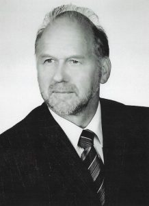 Zdjęcie czarno-białe, portretowe, formalne, wykonane w wysokiej jakości – prawdopodobnie w warunkach studyjnych lub na potrzeby oficjalne (np. do biografii, strony instytucji, publikacji). Przedstawia mężczyznę w ujęciu od klatki piersiowej w górę, ustawionego lekko pod kątem, z twarzą skierowaną delikatnie w lewo (dla widza – w prawo). Mężczyzna patrzy wprost przed siebie lub nieco powyżej obiektywu, jego spojrzenie jest spokojne, życzliwe i pełne powagi.Wygląd mężczyzny:Mężczyzna w wieku około 60–70 lat. Twarz owalna, z wyraźnymi rysami i naturalnymi oznakami starzenia – zmarszczki mimiczne, zwłaszcza wokół oczu i czoła. Broda krótka, równo przystrzyżona, podobnie jak wąsy – zarost jasny, siwawy. Czoło wysokie, częściowo łysiejące – linia włosów cofnięta, ale zachowane pasma włosów są zaczesane do tyłu i na bok. Włosy jasne lub siwe, lekko falowane, zadbane.Oczy jasne (prawdopodobnie niebieskie lub szare – nie do ustalenia ze względu na brak koloru), brwi średnio gęste, lekko zakrzywione. Nos prosty, średniej długości. Usta zamknięte, w lekkim półuśmiechu – wyraz twarzy przyjazny, profesjonalny i pogodny.Ubiór:Mężczyzna ubrany jest bardzo elegancko – ma na sobie ciemny garnitur w delikatny wzór (prawdopodobnie prążki), białą koszulę oraz krawat w ukośne paski o różnej szerokości. Krawat starannie zawiązany, jego wzór kontrastuje subtelnie z garniturem. Całość ubioru sugeruje profesjonalizm i klasę – możliwe, że jest to osoba pełniąca funkcję nauczyciela, dyrektora, naukowca lub urzędnika.Tło i kompozycja:Tło jest jasne, zupełnie jednolite – prawdopodobnie białe lub jasnoszare. Brak jakichkolwiek przedmiotów czy dekoracji – tło neutralne, nieodwracające uwagi od twarzy. Oświetlenie miękkie, równomierne, bardzo dobrze zbalansowane – twarz jest dobrze oświetlona, bez ostrych cieni, co sprawia, że zdjęcie ma charakter oficjalny i estetyczny.Wrażenie ogólne:Portret przedstawia osobę doświadczoną, zaufaną, spokojną i godną szacunku. Styl fotografii oraz ubiór sugerują profesjonalizm, autorytet i życzliwość. Zdjęcie mogło zostać wykonane na potrzeby szkoły, instytucji edukacyjnej, naukowej lub urzędowej.