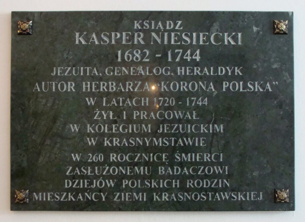 Opis fizyczny tablicy: Tablica jest prostokątna, wykonana z ciemnozielonego lub czarnego marmuru z delikatnym żyłkowaniem. Cztery ozdobne metalowe nity w rogach tablicy przytwierdzają ją do ściany. Tekst wyryty w kamieniu ma srebrny lub biały kolor, czytelny na ciemnym tle. Treść napisu na tablicy: KSIĄDZ KASPER NIESIECKI 1682 – 1744 JEZUITA, GENEALOG, HERALDYK AUTOR HERBARZA „KORONA POLSKA” W LATACH 1720 – 1744 ŻYŁ I PRACOWAŁ W KOLEGIUM JEZUICKIM W KRASNYMSTAWIE W 260 ROCZNICĘ ŚMIERCI ZASŁUŻONEMU BADACZOWI DZIEJÓW POLSKICH RODZIN MIESZKAŃCY ZIEMI KRASNOSTAWSKIEJ Dodatkowe informacje wizualne: Napis wykonany klasyczną, prostą, bezszeryfową czcionką. W górnej i dolnej części tablicy znajdują się małe metalowe rozety (prawdopodobnie mosiężne), użyte jako ozdobne mocowania.