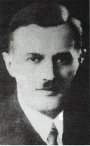 Czarno-białe, lekko rozmazane zdjęcie portretowe przedstawia mężczyznę w średnim wieku, ujętego od klatki piersiowej wzwyż. Jego twarz skierowana jest na wprost, lekko zwrócona w lewo względem widza. Zdjęcie ma dość niską jakość – kontrast i ostrość są ograniczone, przez co detale są lekko zamglone, jednak ogólny wygląd postaci jest rozpoznawalny. Mężczyzna ma regularne rysy twarzy, wyraźnie zarysowane kości policzkowe i prosty nos. Usta są zamknięte, wyraz twarzy poważny, neutralny, bez uśmiechu. Posiada wąsy – cienkie, ale wyraźnie zaznaczone, pokrywające górną wargę. Oczy są ciemne, nieco zasłonięte cieniem, ale patrzą bezpośrednio przed siebie. Włosy są przycięte krótko i uczesane schludnie, zaczesane na bok. Linia włosów jest regularna, z lekkim zakolem po bokach. Włosy są ciemne, a całe czoło gładkie i szerokie. Mężczyzna ubrany jest formalnie: ma na sobie jasną koszulę z kołnierzykiem, krawat oraz ciemną marynarkę. Krawat ma widoczny, ale nieostry wzór. Kołnierzyk koszuli odstaje lekko nad klapami marynarki. Tło zdjęcia jest całkowicie ciemne, jednolite, bez żadnych widocznych elementów, co dodatkowo skupia uwagę na twarzy. Zdjęcie najprawdopodobniej zostało wykonane w pierwszej połowie XX wieku, być może w okresie międzywojennym. Jego styl wskazuje na oficjalny lub dokumentacyjny charakter – może to być fotografia legitymacyjna, urzędowa, bądź wykonana do publikacji prasowej lub archiwum.