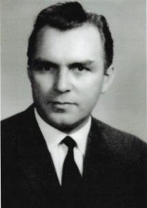 Czarno-białe zdjęcie portretowe, oficjalne, wykonane w stylu studyjnym lub dokumentacyjnym – typowym dla połowy XX wieku. Przedstawia mężczyznę w ujęciu od klatki piersiowej w górę. Postać jest ustawiona frontalnie – mężczyzna patrzy prosto w obiektyw, co nadaje zdjęciu formalny, bezpośredni charakter.
Wygląd mężczyzny:Mężczyzna w wieku około 35–45 lat. Twarz proporcjonalna, o regularnych rysach – czoło wysokie, oczy duże, głęboko osadzone, spojrzenie poważne i skupione. Brwi gęste, naturalnie łukowate. Nos prosty, średniej wielkości. Usta zamknięte, wyraz twarzy neutralny, bez uśmiechu, ale nie surowy. Twarz gładko ogolona, bez zarostu.Włosy ciemne, gęste, zaczesane starannie na bok z lekkim uniesieniem na czubku głowy – typowa fryzura męska z lat 50. i 60. XX wieku. Linia włosów naturalna, lekko cofnięta przy skroniach.
Ubiór:Mężczyzna ubrany jest formalnie – ma na sobie ciemny garnitur (prawdopodobnie czarny lub grafitowy), białą koszulę i czarny krawat. Krój marynarki klasyczny, z wyraźnie zarysowanym kołnierzem. Krawat wąski, dopasowany, typowy dla eleganckiej mody męskiej połowy XX wieku. Cały strój jest schludny i zadbany, co podkreśla powagę i oficjalność portretu.
Tło i oświetlenie:Tło jasne, jednolite, bez elementów dekoracyjnych – najprawdopodobniej biała lub jasnoszara ściana fotograficzna. Oświetlenie miękkie, równomierne, bez ostrych cieni – twarz mężczyzny dobrze doświetlona, co podkreśla rysy bez przerysowań. Zdjęcie wyraźne, ostre, estetycznie zrównoważone.
Wrażenie ogólne:Fotografia przedstawia osobę o nienagannym wyglądzie i postawie, która mogła pełnić funkcję nauczyciela, urzędnika, naukowca lub pracownika instytucji publicznej. Zdjęcie sprawia wrażenie wykonania do dokumentów oficjalnych, książki pamiątkowej lub galerii zasłużonych.