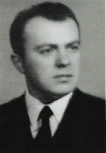 Zdjęcie czarno-białe, portretowe, w stylu formalnym, wykonane prawdopodobnie w połowie XX wieku. Przedstawia mężczyznę sfotografowanego od klatki piersiowej w górę, ustawionego lekko bokiem, z głową skierowaną w lewo (z punktu widzenia widza – w prawo). Mężczyzna nie patrzy bezpośrednio w obiektyw, jego wzrok skierowany jest w dal, poza kadr.Twarz mężczyzny ma wyraz poważny i zdecydowany. Rysy twarzy regularne, czoło wysokie, oczy głęboko osadzone, spojrzenie skupione. Nos prosty, usta zamknięte, wyraz twarzy neutralny – ani uśmiechnięty, ani ponury. Twarz owalna, lekko kwadratowa przy szczęce.Włosy krótkie, ciemne, gęste, zaczesane do tyłu i lekko na bok, z delikatnym przedziałkiem. Linia włosów naturalna, zakola lekko zarysowane. Brak zarostu – twarz ogolona gładko.Mężczyzna ubrany jest bardzo elegancko, w ciemny garnitur, białą koszulę i ciemny krawat. Strój sugeruje, że fotografia została wykonana na potrzeby oficjalne – do dokumentów, pracy zawodowej, książki pamiątkowej lub archiwum. Kołnierzyk koszuli wyraźnie widoczny, starannie ułożony. Marynarka dobrze dopasowana, klasyczna w kroju.Tło zdjęcia jest jednolite, szare lub jasnoszare, bez żadnych wzorów ani przedmiotów. Oświetlenie jest miękkie, równomierne, skierowane z przodu i lekko z boku – twarz mężczyzny dobrze oświetlona, bez ostrych cieni.Cała kompozycja jest stonowana i formalna, bez żadnych ozdób, biżuterii, czy innych rozpraszających elementów. Zdjęcie sugeruje, że przedstawiona osoba była prawdopodobnie nauczycielem, urzędnikiem, działaczem społecznym lub osobą publiczną.