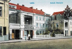 Zdjęcie to przedstawia kolorowaną ilustrację budynku szkoły, wykonaną w stylu starej pocztówki. Budynek zajmuje większą część kadru, widoczny jest od frontu. Scena ma charakter pogodny, dzienny, z wyraźnymi detalami architektonicznymi i historyczną zabudową. Budynek: Główna część to dwupiętrowy budynek z jasną fasadą – parter i piętro mają okna równo rozmieszczone w poziomie. Fasada utrzymana w jasnych kolorach: kremowym, szarym i białym. Dach dwuspadowy, pokryty czerwonymi dachówkami. Po lewej stronie wyróżnia się balkon wsparty na kolumnach, znajdujący się nad wejściem z napisem: „KSIĘGARNIA I SKŁAD GAZET J. CH. HERTZA”. Nad balkonem jest zadaszenie i balustrada. Na parterze widoczne są duże witryny sklepowe i drzwi wejściowe, które mogą należeć do księgarni. Otoczenie: Przed budynkiem znajduje się niski, biały, drewniany płotek i drzewka ozdobne. Na chodniku widoczna jest dwójka ludzi – dorosły mężczyzna w ciemnym fraku i kobieta w jasnej sukni z kapeluszem. Ulica przed budynkiem jest szeroka i pusta, brukowana lub asfaltowa, bez pojazdów.