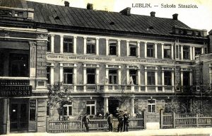 To zdjęcie przedstawia ten sam budynek, ale w bardziej realistycznej i czarno-białej wersji. Ujęcie jest niemal identyczne, lecz zdjęcie wykonano z nieco niższej perspektywy i w innej porze dnia.
Budynek:Główna bryła budynku pozostaje ta sama – trzy kondygnacje (parter i dwa piętra). Elewacja jest bardziej wyrazista dzięki cieniom i światłu, co uwidacznia gzymsy, okna i tynk.Na pierwszym piętrze widoczny jest długi baner z patriotycznym napisem (cytat z epoki z motywem narodowym):
„Naród, który nie zna swej przeszłości, umiera i nie buduje przyszłości.”Po lewej stronie ponownie widzimy wejście do księgarni, z szyldem „KSIĘGARNIA JÓZEF CHOWSKI”.Otoczenie:Przed budynkiem, za drewnianym ogrodzeniem, stoi grupa kilku mężczyzn i chłopców – prawdopodobnie uczniowie lub nauczyciele, ubrani w długie płaszcze, marynarki i czapki z daszkiem.Na parterze, w oknach i drzwiach widoczne są osoby – część spogląda przez okna, co sugeruje aktywność wewnątrz szkoły.Otoczenie wydaje się spokojne, bez ruchu ulicznego – na pierwszym planie widoczna nawierzchnia ulicy.Podsumowanie:Oba obrazy przedstawiają ten sam zabytkowy budynek w Lublinie – dawną Szkołę Staszica, ukazaną w różnych momentach. Budynek posiada cechy klasycznej architektury miejskiej przełomu XIX i XX wieku, z wyraźnymi elementami edukacyjnymi i kulturalnymi (szkoła, księgarnia). Obrazy dokumentują życie codzienne oraz urbanistykę przedwojennego Lublina.