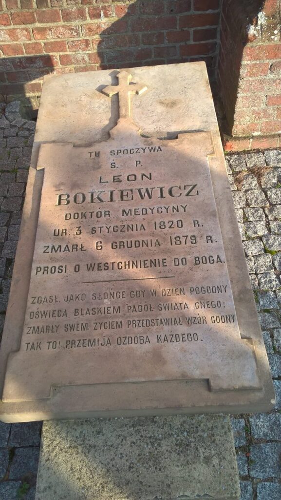 Zdjęcie przedstawia płaską płytę nagrobną z jasnego piaskowca lub kamienia, umieszczoną poziomo, przed ceglanym murem. Płyta znajduje się na chodniku z kostki granitowej. Na nagrobku wygrawerowany jest ozdobny krzyż oraz długi tekst epitafijny. Opis szczegółowy: W górnej części nagrobka, centralnie, wyrzeźbiony jest krzyż łaciński w płaskim reliefie. Poniżej krzyża tekst wykonany jest dużymi, wyraźnymi literami drukowanymi, częściowo pogrubionymi: TU SPOCZYWA Ś. P. LEON BOKIEWICZ DOKTOR MEDYCYNY UR. 3 STYCZNIA 1820 R. ZMARŁ 6 GRUDNIA 1879 R. PROSI O WESTCHNIENIE DO BOGA. Dolna część nagrobka zawiera dłuższy tekst epitafijny – refleksyjny, metaforyczny opis śmierci i życia zmarłego: ZGASŁ JAKO SŁOŃCE GDY W DZIEŃ POGODNY OŚWIECA BLASKIEM PADOŁ ŚWIATA CNEGO; ZMARŁY SWEM ŻYCIEM PRZEDSTAWIAŁ WZÓR GODNY TAK TO! PRZEMIJA OZDOBA KAŻDEGO. Płyta nagrobna nosi ślady starzenia – widoczne są przebarwienia, ubytki i miejscowe odbarwienia od warunków atmosferycznych.