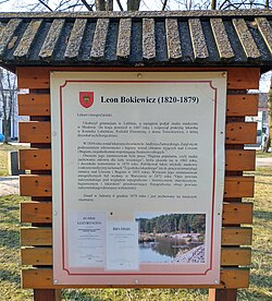 Zdjęcie przedstawia dużą tablicę informacyjną poświęconą Leonowi Bokiewiczowi (1820–1879), ustawioną na tle parku lub skweru. Tablica umieszczona jest w drewnianej, zadaszonej konstrukcji i zawiera biograficzny tekst oraz zdjęcia. Opis szczegółowy: Tablica ma tło jasne, obramowane na czerwono. U góry znajduje się herb miasta oraz czerwony nagłówek: "Leon Bokiewicz (1820–1879)" Poniżej znajduje się dłuższy tekst opisujący: Życie Bokiewicza w Lublinie i jego zasługi jako lekarza. Działalność w dziedzinie zdrowia publicznego. Informacje o jego śmierci i pochówku. W dolnej części tablicy umieszczone są dwa elementy graficzne: Po lewej: skan nekrologu z epoki (mało czytelny na zdjęciu). Po prawej: współczesne zdjęcie przyrody – najprawdopodobniej jeziora lub parku, być może miejsca upamiętnienia. Otoczenie: Zdjęcie wykonano na zewnątrz w pogodny dzień – w tle widoczne są drzewa bez liści oraz trawniki, co sugeruje porę wczesnowiosenną lub późnojesienną.