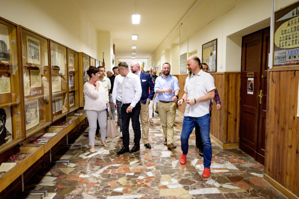 Grupa dorosłych osób (około 12 widocznych) idzie korytarzem szkolnym z charakterystyczną, kolorową mozaikową podłogą. Po lewej stronie znajdują się oszklone gabloty muzealne, zawierające dokumenty, zdjęcia, pamiątki historyczne i plansze z tekstami. Po prawej – drzwi do sal lekcyjnych i wyłożona boazerią ściana. Uczestnicy rozmieszczają się luźno w grupach, rozmawiają, śmieją się, niektórzy patrzą na eksponaty. Atmosfera jest radosna i sentymentalna. Styl ubioru: elegancko-casualowy. Wyróżnia się mężczyzna na pierwszym planie w białej koszuli z krótkim rękawem i czerwonych butach sportowych.