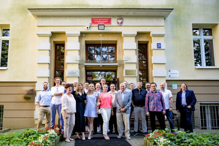 Zdjęcie przedstawia grupę 21 osób stojących przed wejściem głównym do budynku szkolnego – klasycznej fasady z dużymi, drewnianymi drzwiami i tablicą z nazwą szkoły: „I Liceum Ogólnokształcące im. Stanisława Staszica w Lublinie” Nad nią znajduje się godło Polski (biały orzeł), a obok tabliczki informacyjne i numer budynku: 26. Osoby stoją na tle kwiatowych rabatek, na chodniku. Ustawieni są w dwóch rzędach – frontowy i tylny. Stroje półformalne: koszule, bluzki, sukienki, garnitury. Kobiety w sandałach i szpilkach, niektóre z torebkami. Kilka osób ma skrzyżowane ręce, inni przytulają się lekko – atmosfera przyjazna i luźna.