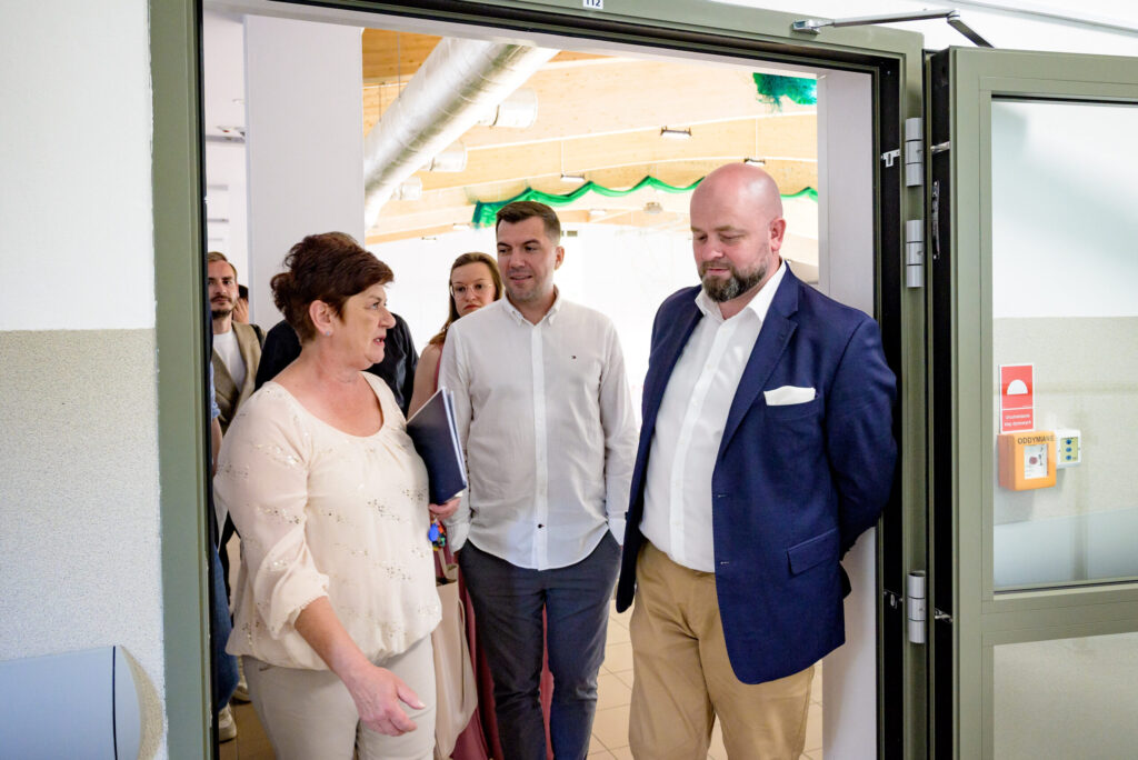 Na zdjęciu widzimy grupę osób stojących w wejściu do nowoczesnej sali szkolnej, której sufit wykończony jest drewnem i metalowymi rurami wentylacyjnymi. Na pierwszym planie kobieta w jasnej bluzce z cekinami prowadzi rozmowę z dwoma mężczyznami – jeden z nich ubrany jest w granatową marynarkę i beżowe spodnie, drugi w białą koszulę. W tle widoczni są inni uczestnicy spotkania – kobiety i mężczyźni w różnym wieku, czekający na wejście do sali. Na ścianie po prawej stronie widnieje przycisk alarmowy i tabliczka z napisem „ODDYMIANIE”.