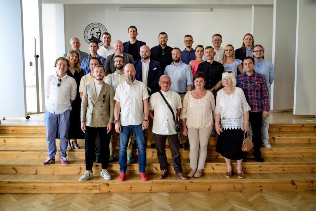 Grupowe zdjęcie wykonane w jasnym pomieszczeniu – najprawdopodobniej na szkolnej scenie lub auli, ustawionej w układzie schodkowym z drewnianych podestów. Na tle białej ściany z logo szkoły („I Liceum Ogólnokształcące im. Stanisława Staszica”) stoi grupa 27 osób – kobiet i mężczyzn w różnym wieku, od około 30 do 70 lat. Osoby rozmieszczone są w trzech rzędach: część stoi, część stoi na wyższych stopniach sceny. Przeważają mężczyźni. Styl ubioru: nieformalny i półformalny. Mężczyźni w koszulach, marynarkach, część z brodami, kobiety w letnich bluzkach, sukienkach, jedna w jasnej koronkowej bluzce. Panuje pozytywna atmosfera – osoby uśmiechają się, stoją blisko siebie.