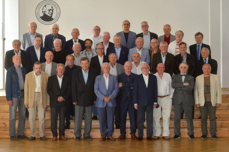 Zdjęcie przedstawia dużą grupę około 36 starszych mężczyzn, stojących razem do wspólnego zdjęcia pamiątkowego na tle białej ściany. Tło to charakterystyczne miejsce: ściana z czarnym, okrągłym logo szkoły zawierającym profil mężczyzny i napis: „I LICEUM OGÓLNOKSZTAŁCĄCE IM. STANISŁAWA STASZICA W LUBLINIE” Mężczyźni ustawieni są w czterech rzędach – ci z przodu stoją na podłodze, wyżsi lub młodsi w tylnych rzędach – na stopniach drewnianej sceny. Wszyscy ubrani są elegancko, większość ma na sobie garnitury, marynarki lub koszule, kilka osób nosi muszki lub okulary. Dominują siwe włosy, łysiny, brody – sugerując zaawansowany wiek uczestników, wielu z nich to zapewne absolwenci z kilkudziesięcioletnim stażem od ukończenia szkoły. Wszyscy patrzą w stronę aparatu, niektórzy z lekkimi uśmiechami, atmosfera formalna i pamiątkowa.