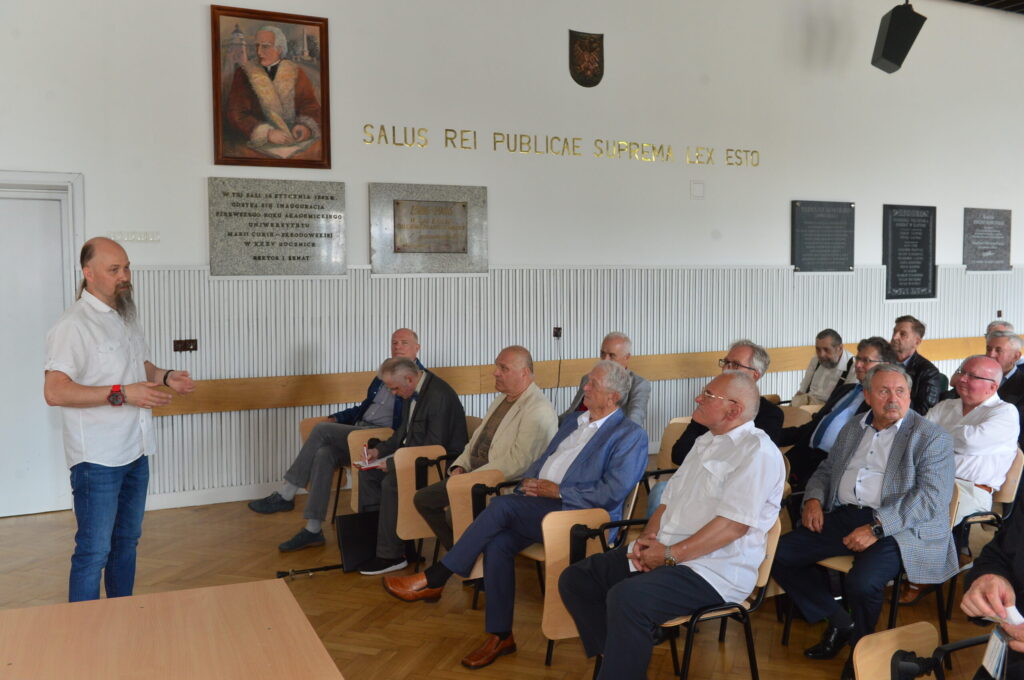 To zdjęcie ukazuje ten sam moment i to samo pomieszczenie, ale z innej perspektywy – bocznej, bliżej portretu i tablic pamiątkowych. Mężczyzna z brodą i długimi włosami (prowadzący) znajduje się po lewej stronie, zwrócony w stronę siedzących uczestników. Większość mężczyzn patrzy uważnie na prowadzącego, niektórzy notują, inni siedzą z założonymi rękami lub rozmawiają cicho. Widać wyraźniej portret na ścianie oraz kamienne tablice: Jedna z napisem: „W tej sali 27 stycznia 1968 r. odbyła się inauguracja pierwszego roku akademickiego Uniwersytetu Marii Curie-Skłodowskiej w Lublinie – filia w Rzeszowie.” Napis łaciński z pozłacanych liter znajduje się wysoko na ścianie i przebiega niemal przez całą długość pomieszczenia.