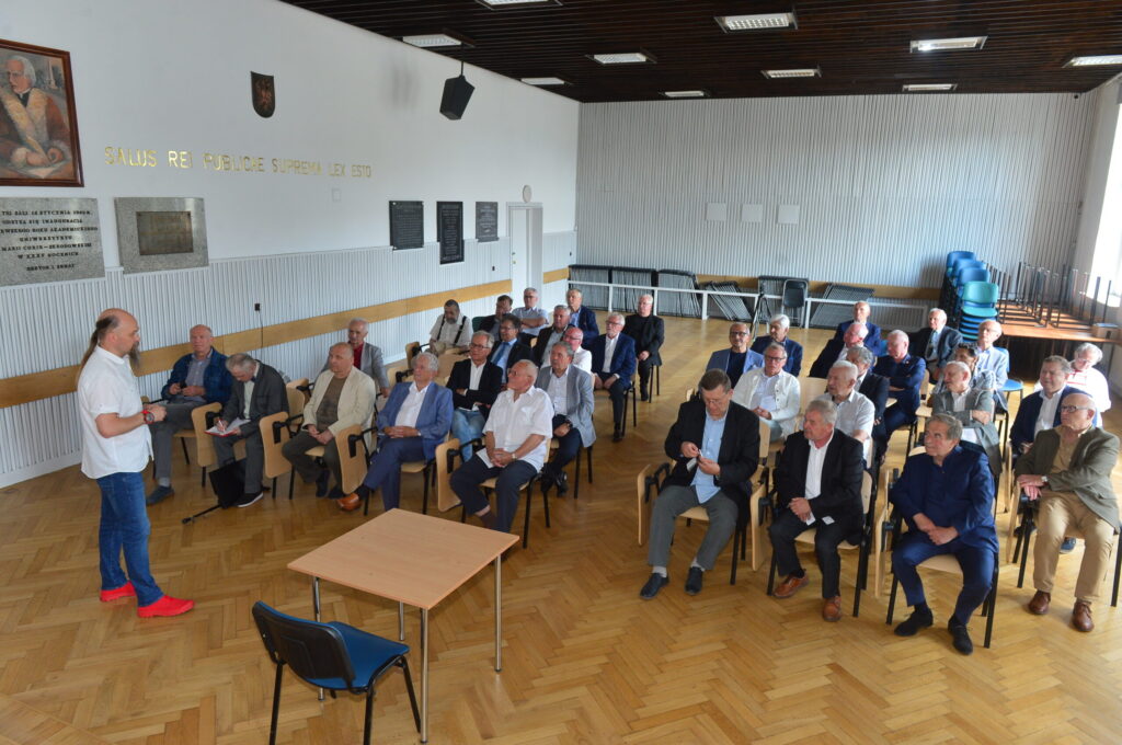 Zdjęcie ukazuje wnętrze szkolnej auli o jasnych ścianach i drewnianym parkiecie w jodełkę, gdzie odbywa się wykład lub prezentacja. W pomieszczeniu znajduje się około 35 mężczyzn, głównie w starszym wieku (60–80 lat), siedzących na krzesłach z jasnym drewnianym oparciem i czarnymi podłokietnikami. Mężczyźni są elegancko ubrani – dominują garnitury, marynarki, koszule, część w dżinsach lub spodniach materiałowych. Kilku z nich ma okulary. Na pierwszym planie, po lewej stronie, stoi mężczyzna z długimi włosami i brodą, w białej koszuli z krótkim rękawem, dżinsach i charakterystycznych czerwonych butach sportowych – prawdopodobnie prowadzący spotkanie. Przed prowadzącym znajduje się niewielki stół oraz puste krzesło z niebieskim siedziskiem. Tło i otoczenie: Na ścianie po lewej stronie widnieje duży portret starszego mężczyzny w historycznym stroju (najpewniej patrona szkoły), herb oraz złoty łaciński napis: „SALUS REI PUBLICAE SUPREMA LEX ESTO” (tłum. „Dobro Rzeczypospolitej niech będzie najwyższym prawem”). Poniżej znajdują się kamienne tablice pamiątkowe, a dalej – czarne tablice z nazwiskami. Z tyłu auli – puste krzesła, złożone stoły, naturalne światło wpadające przez okna (po prawej, poza kadrem).