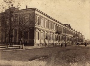 Zdjęcie przedstawia duży, masywny budynek murowany, sfotografowany pod kątem, z narożnika dwóch przecinających się ulic. Budynek rozciąga się wzdłuż horyzontu, pokazując swoją ogromną długość. Fotografia wykonana jest w sepii – odcieniach brązu, typowych dla starych zdjęć z końca XIX lub początku XX wieku. Architektura: Budynek ma dwie kondygnacje (parter i piętro) oraz wysoką piwnicę lub cokół. Ściany wykonane są z czerwonej cegły, w dolnej części wyraźnie ciemniejszej niż górna część elewacji, co może sugerować użycie kamienia lub tynku na cokole. Okna duże, prostokątne, ustawione rytmicznie – na każdym piętrze tworzą długi ciąg identycznych otworów z drobnym detalem: okna parterowe są prostsze, a nad nimi – okna piętra – mają ozdobne nadproża w formie gzymsów. W połowie długości budynku widoczny jest niewielki, wystający daszek nad wejściem, prawdopodobnie głównym – wskazuje to na drzwi wejściowe, choć są częściowo przysłonięte cieniem. Otoczenie: Przed budynkiem znajduje się brukowana ulica z torami kołowymi i wyboistą powierzchnią. Na skraju chodnika rośnie szpaler drzew bez liści, co sugeruje późną jesień lub zimę. Wzdłuż ulicy ustawiono drewiane ogrodzenia i słupki, niskie, oddzielające ruch pieszy od jezdni. Po lewej stronie, przy narożniku budynku, stoi kilka osób w ciemnych ubraniach – są mało wyraźne, prawdopodobnie uczniowie lub przechodnie. W dalszej części ulicy widoczna jest grupa ludzi, niektórzy idą, inni stoją – postacie są bardzo małe z racji oddalenia. Klimat zdjęcia: Zdjęcie ma melancholijny, spokojny charakter – pustawa ulica, bez pojazdów, szare niebo, brak liści na drzewach. Brak współczesnych elementów infrastruktury (latarnie, znaki) podkreśla historyczny charakter fotografii. Budynek wygląda na instytucję publiczną – prawdopodobnie szkołę, gimnazjum lub urzędowy gmach administracyjny, z uwagi na rozmiar, symetrię i brak ozdobnych detali prywatnej rezydencji. Podsumowanie: Zdjęcie ukazuje monumentalny budynek szkolny lub instytucjonalny w historycznym Lublinie. Ujęcie z rogu ulicy podkreśla jego długość i znaczenie w strukturze miejskiej. Obecność ludzi i szpaleru drzew wskazuje na żyjące otoczenie, a całość zdjęcia wpisuje się w dokumentację miejskiej architektury przełomu XIX i XX wieku.