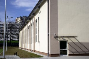 Pokazuje boczną ścianę nowoczesnej sali gimnastycznej z jasnym, gładkim tynkiem (kremowo-beżowym).Elewacja jest rytmicznie podzielona przez wysokie, wąskie okna rozmieszczone pionowo w równych odstępach.Na pierwszym planie znajduje się niskie wejście z przezroczystym daszkiem – proste drzwi z klamką, bez schodów, prowadzące wprost z chodnika.Po lewej stronie widoczna ławka miejska i słup oświetleniowy.W tle, za budynkiem, znajdują się wysokie bloki mieszkalne – co sugeruje miejskie, gęsto zabudowane otoczenie szkoły.