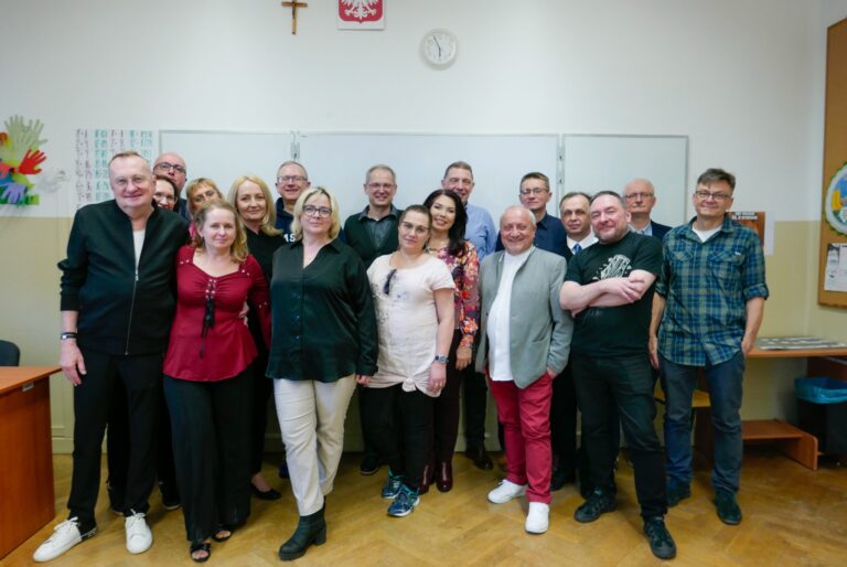 Grupowe zdjęcie 20 osób (kobiet i mężczyzn) wykonane w sali lekcyjnej. Osoby stoją w dwóch rzędach, patrząc w obiektyw i uśmiechając się. Tło to biała tablica, zegar, godło Polski i krzyż. Styl ubioru uczestników: luźno-elegancki. Kobiety w ciemnych bluzkach, jedna w czerwonej sukience, mężczyźni w koszulach, swetrach lub T-shirtach.