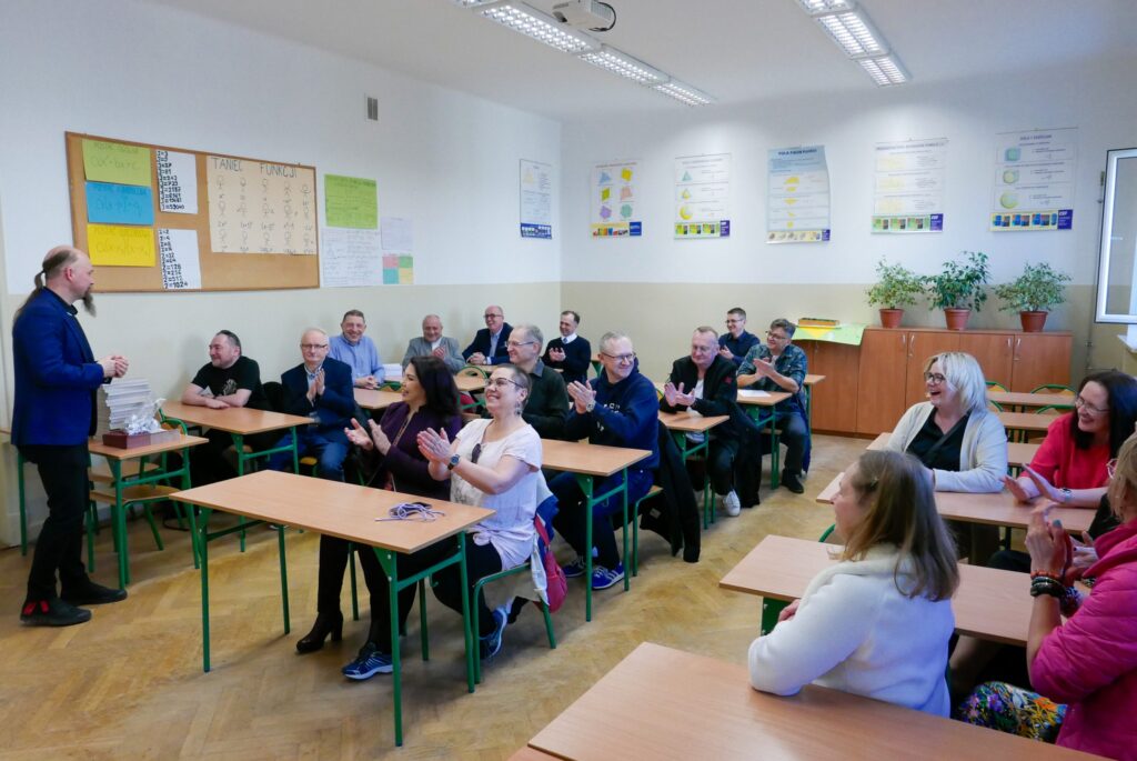 Zdjęcie przedstawia nowoczesną klasę lekcyjną z jasnym światłem sufitowym. Przy szkolnych ławkach z drewnianymi blatami i zielonymi metalowymi nogami siedzi około 20 dorosłych osób – kobiety i mężczyźni, w większości w wieku 40–60 lat. Wszyscy patrzą w stronę prowadzącego – mężczyzny w granatowej marynarce i z długą brodą, który stoi przy biurku nauczyciela. Na ścianach wiszą plakaty edukacyjne z geometrią, a na dużej tablicy korkowej widoczny jest rysunek zatytułowany „Taniec funkcji” – z rysunkowymi ludzikiem przedstawiającym wykres funkcji sinus.