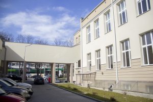 Zdjęcie przedstawia zewnętrzną część szkoły – fragment elewacji głównego budynku z jasnym tynkiem i dużymi, białymi oknami. Po lewej stronie widoczny jest parking szkolny – kilka zaparkowanych samochodów. Nad częścią przejazdu znajduje się łącznik – zabudowany korytarz biegnący nad otwartym przejściem, łączący dwa skrzydła budynku. Łącznik ma prostą, jasną formę z płaskim dachem. W tle, przez otwarte przejście, widać kolejne zabudowania oraz fragmenty hal sportowych i boisk.