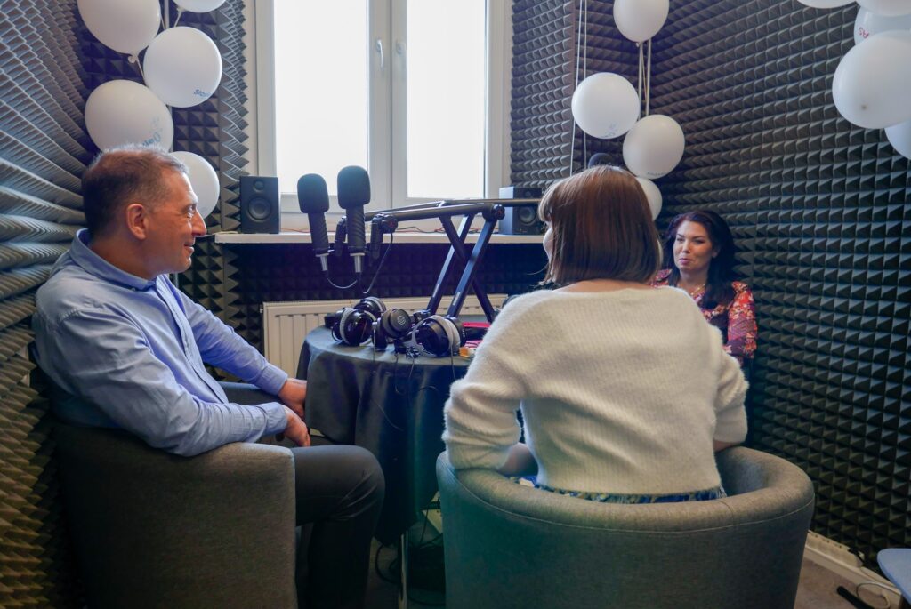 To samo studio co wcześniej, ale widziane z perspektywy okna. Widać trzy osoby: kobieta tyłem, mężczyzna w błękitnej koszuli, oraz kobieta w kwiatowej bluzce. Wszyscy prowadzą rozmowę. Na parapecie białe głośniki, na ścianach panele akustyczne i balony. Na stole leżą słuchawki i notatki.