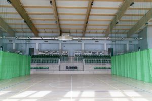 Obiekt sfotografowany z poziomu boiska – widok centralny, prosto na trybuny. Podłoga hali to jasne, sportowe, syntetyczne tworzywo z wyraźnie widocznymi liniami boiskowymi (białe linie: koło środkowe i linie boczne). Po obu stronach zdjęcia rozwieszone są zielone zasłony z siatki – służące do dzielenia przestrzeni hali na części treningowe. Na końcu hali znajduje się wysoka trybuna z zielonymi, składanymi siedzeniami i dwiema kabinami komentatorskimi lub stanowiskami technicznymi nad siedzeniami. Sufit wykonany z drewnianych belek poprzecznych i podwieszonego oświetlenia – nowoczesna, jasna przestrzeń z dobrą akustyką.