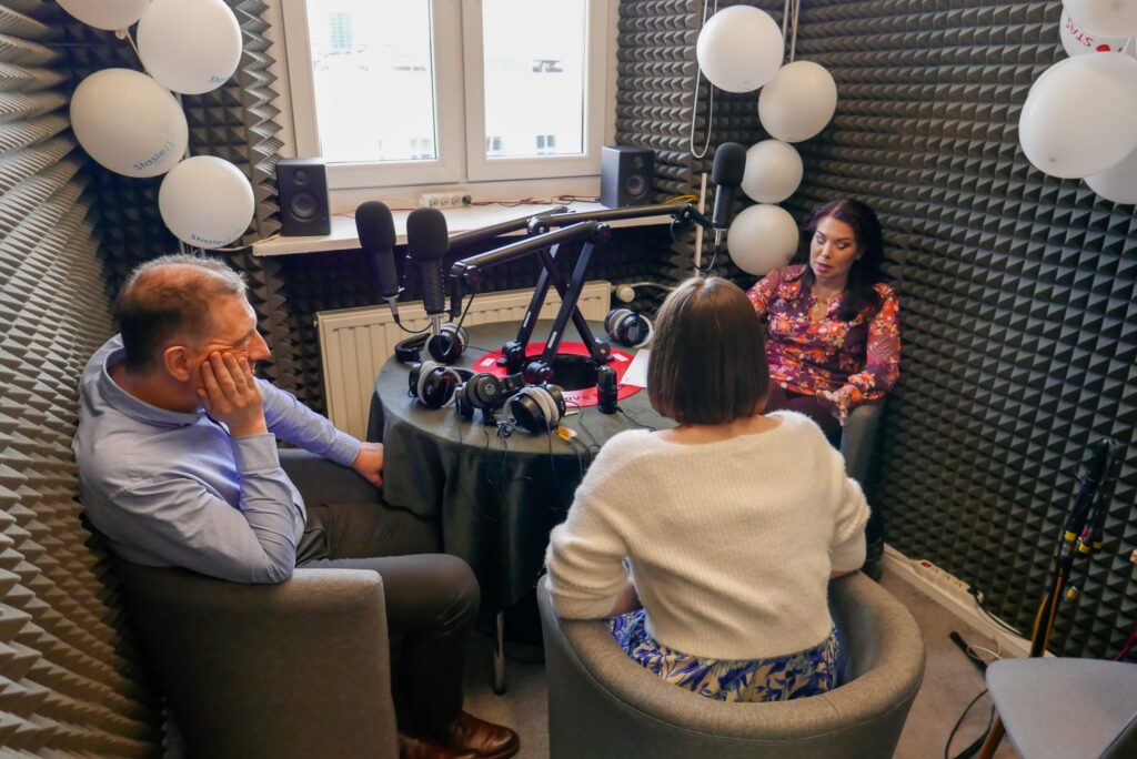 Wygłuszone studio radiowe z czarnymi piankami akustycznymi. Trzy osoby (dwie kobiety i jeden mężczyzna) siedzą wokół małego, okrągłego stołu. Na środku znajdują się cztery mikrofony na ramionach i kilka zestawów słuchawkowych. Na ścianie białe balony z logo „Stasie Radio”. Kobieta w kolorowej bluzce mówi, pozostałe osoby słuchają.