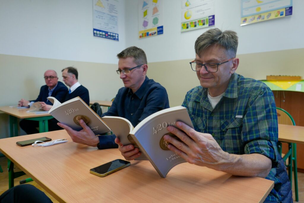 Dwóch mężczyzn siedzi przy ławce i czyta książkę zatytułowaną „420 lat Lubelskiego Staszica” z medalionem na okładce. Mają skupiony wyraz twarzy. Na ławce obok leżą smartfony, okulary, długopis i słuchawki. W tle kolejne osoby oraz geometryczne plansze edukacyjne na ścianie.