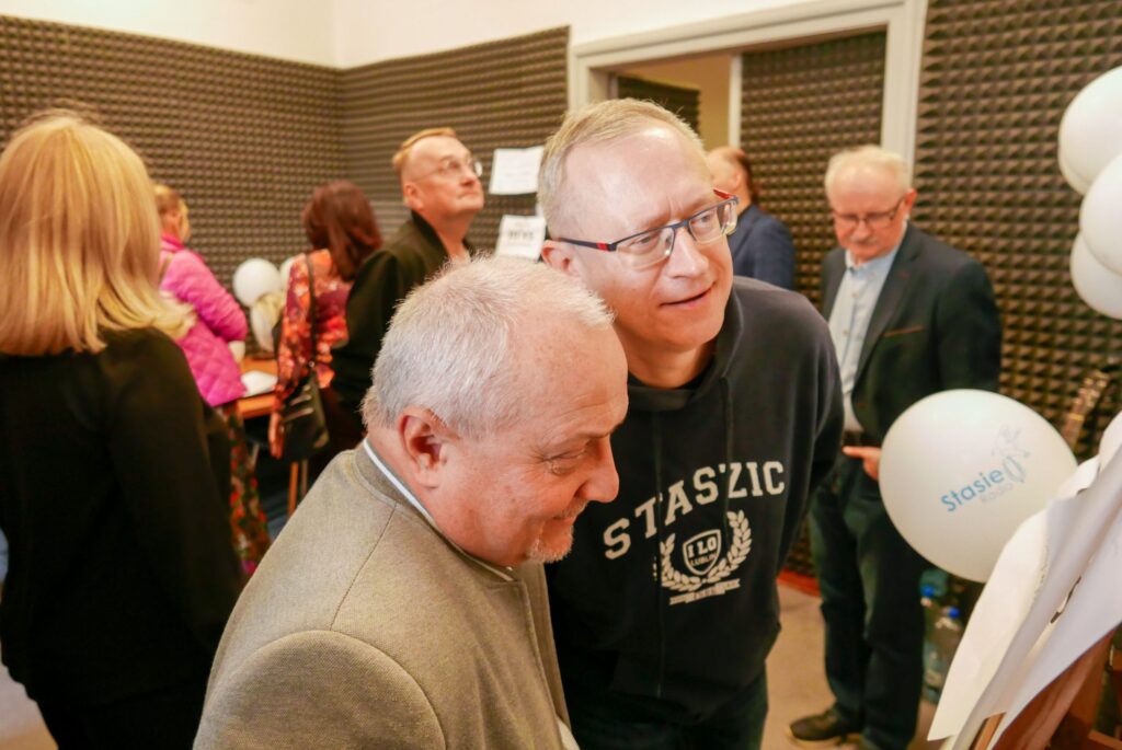 Wnętrze ciasnego, wygłuszonego pomieszczenia przypominającego studio nagrań. Dwa mężczyźni stoją blisko siebie i pochylają się nad dużą tablicą lub planszą. Jeden z nich ma na sobie bluzę z napisem „STASZIC I LO Lublin”, drugi – jasnoszarą marynarkę. W tle inne osoby i białe balony z napisem „Stasie Radio”.