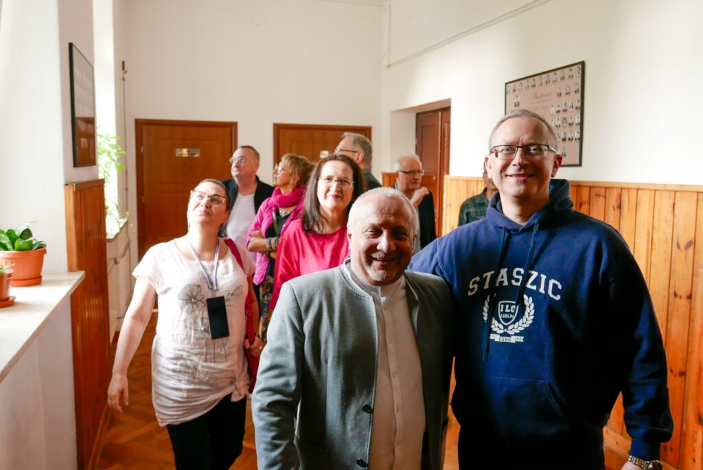 Grupa około 12 osób spaceruje jasnym szkolnym korytarzem z drewnianą boazerią. Na pierwszym planie kobieta w jasnoróżowej bluzce z telefonem na smyczy, patrzy w górę. Z tyłu mężczyzna w ciemnej bluzie z napisem „STASZIC” oraz inne osoby ubrane kolorowo, w tym kobieta w fuksjowym szalu. Na ścianie widać tablicę pamiątkową ze zdjęciami dawnych klas.