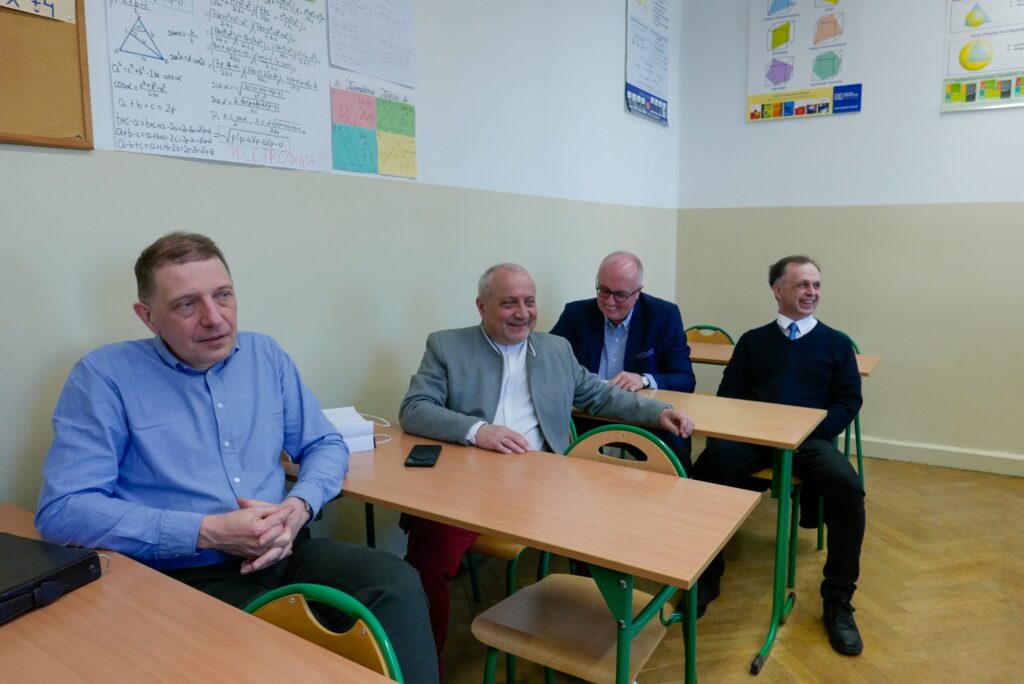 Zbliżenie na czterech mężczyzn siedzących przy szkolnych ławkach. Rozmawiają i śmieją się, panuje luźna atmosfera. Na ścianie nad nimi wiszą ręcznie robione plansze z wzorami geometrycznymi: twierdzenie Pitagorasa, pola trójkątów, wzory skróconego mnożenia. Dwóch mężczyzn nosi marynarki, jeden sweter, jeden błękitną koszulę.