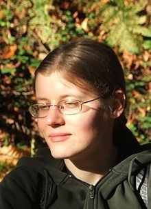 Na zdjęciu widoczna jest młoda kobieta sfotografowana w naturalnym, leśnym otoczeniu. Kadr obejmuje jej twarz i górną część torsu, pokazując ją z profilu lekko odwróconego w lewą stronę (z perspektywy widza). Kobieta ma jasną cerę i średniej długości, jasnobrązowe włosy, starannie zaczesane do tyłu i związane. Ma na sobie okulary z cienkimi, metalowymi oprawkami w kolorze srebrnym. Jej oczy są przymknięte, jakby chroniła się przed światłem lub była pogrążona w spokojnej refleksji. Twarz kobiety oświetlona jest ciepłym, złocistym światłem słońca, co tworzy wyraźny kontrast z ciemniejszymi partiami jej ubioru i tła. Wyraz jej twarzy jest łagodny i spokojny, z lekkim, niemal niezauważalnym uśmiechem. Ubrana jest w ciemnozieloną bluzę z zamkiem błyskawicznym, której kołnierz jest lekko uniesiony. Na jednym ramieniu ma ciemny plecak, widoczny tylko częściowo. Tło stanowi rozmyta roślinność leśna — liście, paprocie i gałęzie — w odcieniach zieleni, brązu i złota. Światło słoneczne przenika przez liście, tworząc jasne plamy i cienie na tle i na twarzy kobiety. Cały obraz emanuje spokojem, ciszą i kontemplacją, jakby przedstawiona osoba znajdowała się w chwili wyciszenia podczas spaceru po lesie w jesienne popołudnie.