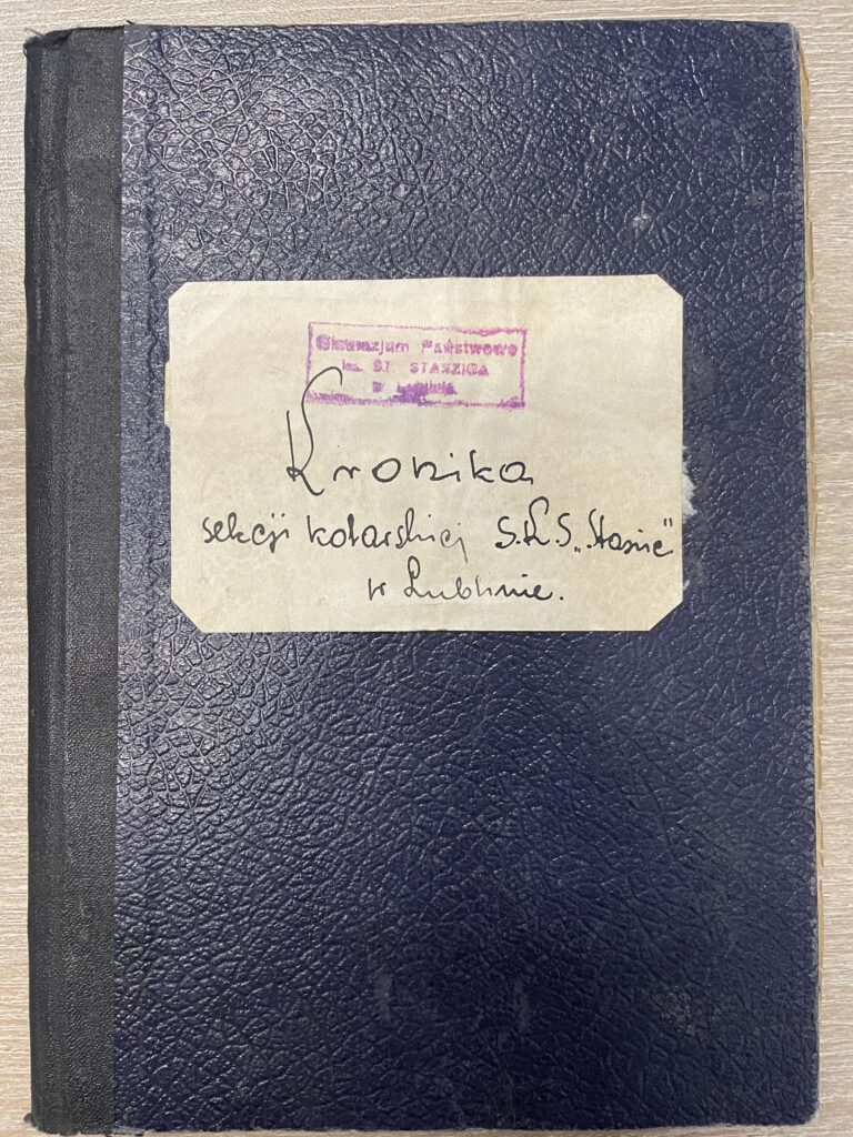 Na zdjęciu widoczna jest zamknięta, stara kronika w formie zeszytu lub skoroszytu. Obiekt leży poziomo na jasnobrązowym, drewnianym blacie. Opis fizyczny zeszytu: Okładka ma ciemnogranatowy, niemal czarny kolor o fakturze przypominającej skórę – z widoczną strukturą pęknięć i linii, nadającą jej stary i zużyty wygląd. Grzbiet zeszytu jest czarny, wykonany z materiału przypominającego taśmę introligatorską, który może być z tkaniny lub innego mocnego materiału. Okładka jest nieco zniszczona – szczególnie przy narożnikach, gdzie widać lekkie przetarcia. Opis naklejki z treścią: Na środku okładki znajduje się prostokątna, pożółkła etykieta przyklejona na stałe. Rogi etykiety są ścięte pod kątem 45 stopni (szczególnie widoczne górne i dolne rogi po prawej stronie). Na etykiecie znajduje się tekst ręczny zapisany piórem lub cienkopisem oraz pieczątka w kolorze fioletowym. Treść etykiety: Na górze, lekko wyblakła pieczątka w kolorze fioletowym: Gimnazjum Państwowe ST. STASZICA w Lublinie – Pieczątka jest lekko przekrzywiona w lewo i nieco rozmazana. Pod pieczątką, większym, ozdobnym pismem odręcznym (kaligrafia) znajduje się tytuł: „Kronika” Niżej, mniejszym pismem odręcznym (bardziej pochyłym), znajduje się opis: „sekcji kolarskiej S.H.S. „Staszic” w Lublinie.” – Skrót S.H.S. prawdopodobnie oznacza „Szkolne Hufce Sportowe”. Podsumowanie: Zdjęcie przedstawia kronikę sekcji kolarskiej działającej przy Gimnazjum Państwowym im. Stanisława Staszica w Lublinie, prowadzoną najprawdopodobniej w okresie przedwojennym lub tuż po wojnie. Dokument ten ma charakter archiwalny i może zawierać relacje z wydarzeń sportowych, wycieczek rowerowych oraz działalności uczniów w ramach tej sekcji. Całość wykonana jest w stylu charakterystycznym dla ręcznie pisanych kronik szkolnych z XX wieku.