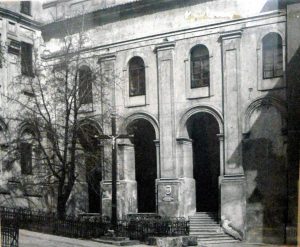 Czarno-białe zdjęcie przedstawia fragment elewacji starego budynku o cechach klasycystycznych, prawdopodobnie z XIX wieku lub wcześniej. Budynek jest wysoki, wielokondygnacyjny, wykonany z kamienia lub tynku o wyraźnie zużytej powierzchni. Zdjęcie jest historyczne, o lekkiej ziarnistości, co sugeruje jego wiek lub pochodzenie z archiwum. Architektura: Budynek posiada wysokie arkadowe wejście z trzema dużymi łukowymi otworami, z których środkowy prowadzi na schody – trzy do pięciu stopni w górę. Fasada ma pionowe zdobienia w postaci pilastrów (płaskie kolumny przylegające do ściany), oddzielające łukowe wnęki. Nad każdym z trzech wejść znajduje się prostokątne okno z łukiem u góry, umieszczone w wysokich, pionowych niszach, co nadaje budowli monumentalny charakter. Tynk na ścianach jest w złym stanie, z widocznymi śladami zabrudzeń, ubytków i łuszczącej się powierzchni. Na ścianie w centralnej części widoczny jest numer 9 umieszczony nad jednym z wejść – prawdopodobnie numer domu. Otoczenie: Po lewej stronie znajduje się drzewo bez liści, które częściowo przysłania fragment elewacji – może to sugerować porę późnej jesieni, zimy lub wczesnej wiosny. Na dole po lewej widoczny jest niski metalowy płot z ozdobnymi pionowymi prętami, który oddziela chodnik od budynku. W dolnej części fotografii widoczna jest ulica lub chodnik – gładki, biegnący wzdłuż budynku. Światło i cień: Oświetlenie jest naturalne, światło słoneczne pada z prawej strony, rzucając wyraźny cień budynku na elewację oraz na część ulicy. Cienie są głębokie, co sugeruje mocne światło słoneczne i kontrast między oświetlonymi a zacienionymi powierzchniami. Ogólna atmosfera: Zdjęcie ma melancholijny, historyczny charakter – przedstawia zaniedbaną, ale monumentalną architekturę miejską z przeszłości. Może przedstawiać dawną szkołę, klasztor, budynek urzędowy lub instytucję publiczną, obecnie nieco zapomnianą. Styl i uszkodzenia mogą sugerować również budynek po wojnie lub okresie opuszczenia.