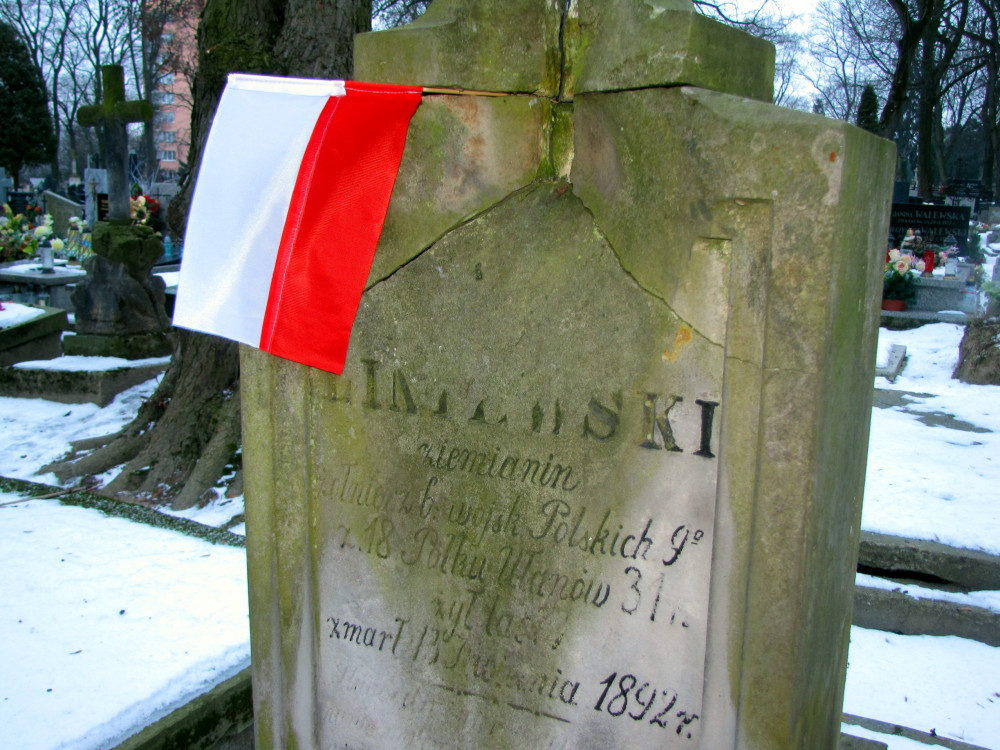 Opis ogólny:
Zdjęcie przedstawia stary, mocno zwietrzały i popękany kamienny nagrobek na cmentarzu, z zawieszoną na nim niewielką polską flagą (biało-czerwoną). Scena ma miejsce zimą – na ziemi i grobach wokół leży śnieg. W tle widać inne groby, krzyże, kwiaty i drzewa bez liści.Opis szczegółowy:Nagrobek wykonany z jasnoszarego piaskowca, ma formę prostego, prostokątnego pionowego pomnika z trapezowym zakończeniem u góry.Płyta jest silnie spękana – głęboka szczelina biegnie przez środek pionowo i poziomo, dzieląc górną część na kilka bloków.Na nagrobku wygrawerowany jest napis. Litery są częściowo starte, ale wciąż częściowo czytelne. Wyróżniają się wyraźnie ciemniejsze litery nazwiska „KWIATKOWSKI” (widoczna tylko część).Pod nazwiskiem można dostrzec fragmenty tekstu, który mówi o służbie wojskowej zmarłego (m.in. „uczestnik Wojen Polskich”, „18 Pułku Ułanów”, „zmarł 13 kwietnia 1892 r.”).Na lewym górnym rogu nagrobka zawieszona jest mała, materiałowa flaga Polski – składająca się z dwóch poziomych pasów: białego na górze i czerwonego na dole. Flaga nie jest w pełni rozwinięta, zwisa lekko po skosie, przypięta do krawędzi nagrobka.W tle znajdują się inne nagrobki, w tym duży kamienny krzyż po lewej stronie, a także groby ozdobione kwiatami sztucznymi lub wieńcami. Cmentarz wygląda na zaniedbany, wiele nagrobków jest starych, część pochylona lub zniszczona.Otoczenie:Widoczna zima: cienka warstwa śniegu pokrywa ziemię i część grobów.W tle rosną gołe, wysokie drzewa, bez liści.W oddali po lewej stronie majaczy pomarańczowy budynek (blok mieszkalny lub kamienica), co sugeruje, że cmentarz znajduje się w mieście.Scena jest spokojna, melancholijna – typowa dla historycznych, zimowych ujęć z cmentarzy.