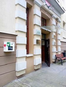 Zbliżenie na główne wejście do budynku. Drzwi drewniane, otwarte, z górnym przeszkleniem. Elewacja jasna, z wyraźnie zaznaczonymi narożnikami wokół drzwi – ozdobne białe prostokątne boniowanie (imitacja kamiennych bloków). Nad drzwiami zamontowana jest przezroczysta osłona przeciwsłoneczna. Na ścianie po lewej znajduje się biała skrzynka z defibrylatorem AED, oznaczona czerwonym sercem i zielonym krzyżem. Obok wejścia stoi drewniana ławka na metalowych nogach. Ściana zawiera także tablice informacyjne i godło szkoły.