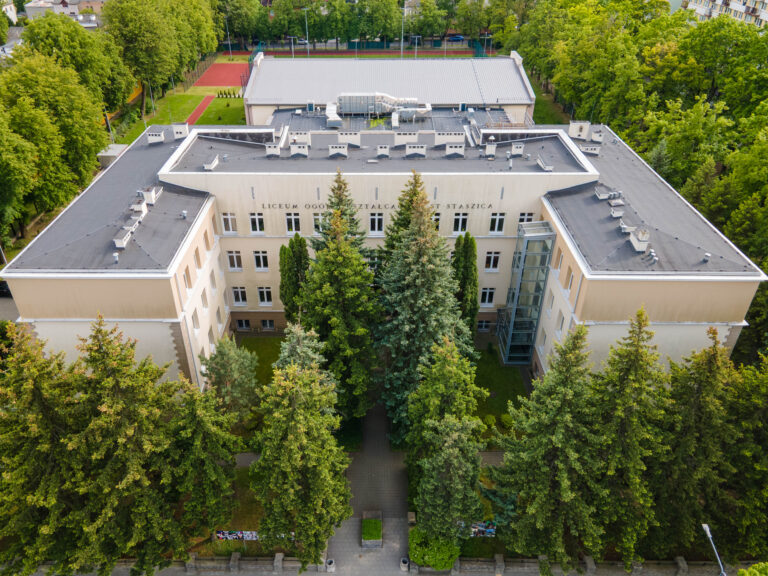 Zdjęcie wykonane z dużej wysokości, pokazuje pełny gmach szkoły od frontu, z dronowej perspektywy. Budynek ma kształt litery U, z trzema głównymi skrzydłami. Elewacja jest jasnobeżowa z jasnymi obramowaniami wokół okien. Dach jest płaski, ciemnoszary. Nad głównym wejściem, w centralnym skrzydle, widoczny jest duży napis: „Liceum Ogólnokształcące im. Staszica”. Przed budynkiem rośnie szpaler gęstych, wysokich świerków i sosen, które częściowo zasłaniają parter. W tle, za budynkiem, widoczne są boiska sportowe z czerwonymi i zielonymi nawierzchniami oraz budynek sali gimnastycznej. Całość otoczona jest gęstą zielenią drzew liściastych – szkoła znajduje się w zadrzewionym terenie miejskim.