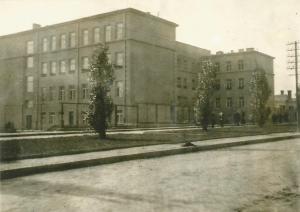 Przedstawia ukończony budynek szkoły – trzy kondygnacje, tynkowana fasada, duże prostokątne okna.Przed budynkiem rosną symetrycznie rozmieszczone młode drzewa liściaste (prawdopodobnie topole lub lipy).Ulica brukowana lub asfaltowa, z chodnikiem i latarnią z prawej strony.Budynek wygląda nowocześnie i czysto – zdjęcie prawdopodobnie z lat 50-60 XX w., tuż po odbudowie lub renowacji.