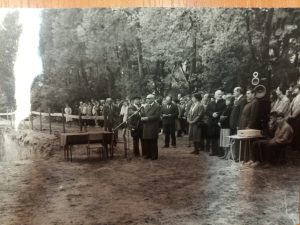 Miejsce wykopu otoczone jest przez duży tłum ludzi – dzieci, nauczycieli i oficjeli. W centrum ustawione są stoły i krzesła, na których leży mikrofon i dokumenty. Widoczne są przemówienia, a w tle drzewa liściaste – miejsce znajduje się w zalesionym terenie.