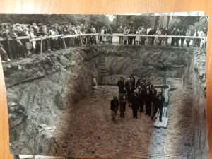 W głębokim wykopie znajduje się grupa około 10 dorosłych mężczyzn w garniturach, w tym duchowny. Wokół wykopu zebrał się tłum uczniów lub mieszkańców, stojących za ogrodzeniem. W centrum wykopu znajduje się symboliczny krzyż i przygotowane miejsce na akt erekcyjny – uroczystość poświęcenia fundamentów.