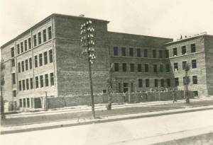 Trzyczęściowy budynek z surowej cegły, bez tynku i detali architektonicznych. Okna już wstawione, ale teren wokół to plac budowy – brak ogrodzenia, tylko niski drut. Widoczna jest słupowa linia energetyczna z dużą liczbą izolatorów.