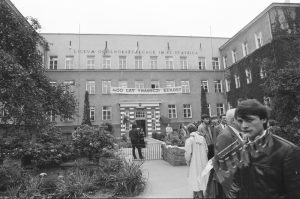 To samo wejście do budynku co wyżej, ale ozdobione napisem na transparencie: „400 lat tradycji szkolnej”. Przed wejściem rozciąga się ogród kwiatowy, uczniowie i goście idą alejką w stronę drzwi. Ludzie ubrani są w kurtki i płaszcze, co sugeruje chłodniejszą pogodę. Na pierwszym planie młody mężczyzna z instrumentem muzycznym (prawdopodobnie trąbka), skierowany w stronę fotografa.