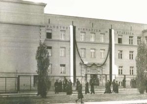 Budynek z prostą elewacją, symetryczny, trzy piętra, płaski dach. Nad wejściem duży napis: „Państwowe Gimnazjum im. Staszica”. Widoczna żałobna dekoracja – czarne wstęgi zawieszone od dachu do wejścia, co sugeruje uroczystość żałobną lub żałobę narodową. Przed szkołą grupa uczniów i dorosłych, ubrane na ciemno.