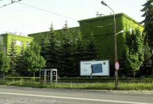 Widok na nowoczesny, prostokątny budynek szkoły, którego elewacja frontowa i boczne ściany są całkowicie porośnięte zielonym bluszczem – tworzy to efekt naturalnej, zielonej ściany. Przed budynkiem rośnie szpaler wysokich świerków, a na chodniku stoi tablica informacyjna z napisem "Zespół Szkół im. Staszica". Wzdłuż ulicy widać ogrodzenie z metalowych prętów, a także latarnię i znaki drogowe. Scena jest zielona, letnia.