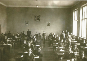 Przedstawia salę lekcyjną z lat 30-40 XX w. – prostokątne pomieszczenie z wysokim sufitem i dużymi oknami po prawej stronie.W klasie siedzi około 30 uczniów w ławkach – chłopcy w ciemnych marynarkach, większość z rękami na blatach.W tle stoi kilku nauczycieli, a na ścianie wiszą portrety (prawdopodobnie narodowych patronów, w tym Stanisława Staszica w centrum).Całość bardzo uporządkowana – uczniowie patrzą w stronę aparatu.
